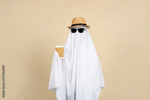 Person in a white ghost costume wearing sunglasses and a straw hat holding a cup of hot coffee on a beige background.