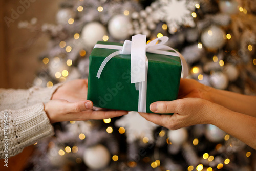 Two hands exchanging a beautifully wrapped green gift box with a white ribbon in front of a decorated Christmas tree with soft glowing lights creating a festive atmosphere