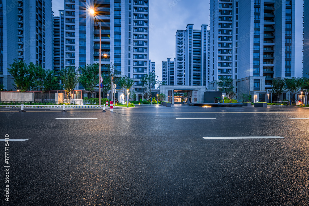 Fototapeta premium Empty wet asphalt road in front of a modern residential apartment complex illuminated by street lights in the evening.