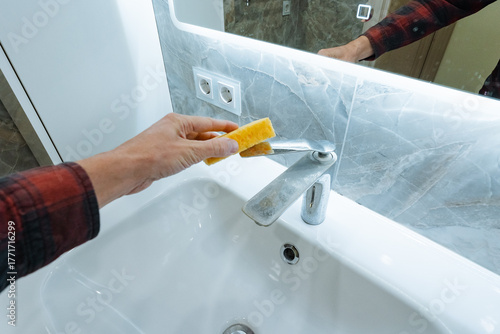 hand wiping sink edge with yellow sponge during bathroom refit, plaid sleeve indicates worker, exposed cabinetry and tools out of frame, focus on grout and seal, practical jobsite