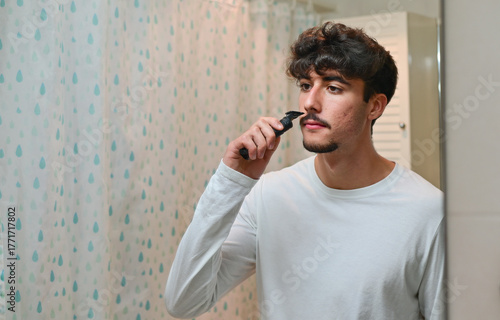 attractive young man shaving beard with electric shaver while looking at mirror in bathroom