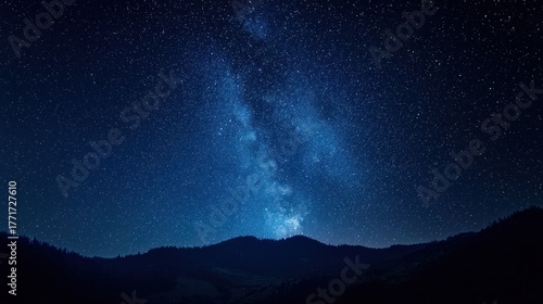 A starry night sky with the Milky Way visible in the distance, silhouetted against the dark blue sky, with mountains in the foreground and a forested area in the background.