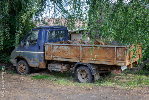 Rusty old blue pickup truck loaded with firewood, parked under leafy trees in a rural setting — perfect for themes of decay, nostalgia, countryside life, and industrial textures.