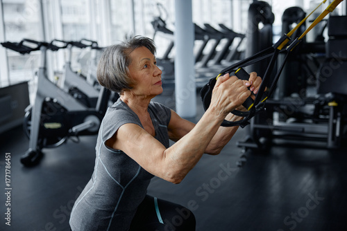 An elderly woman is exercising on a suspension trainer in a gym