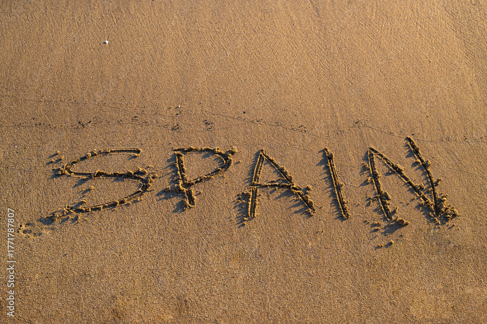 Fototapeta premium The word “Spain” written on sandy beach near the shoreline