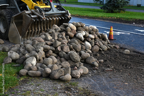 A front loader stands on a wet city road beside a bicycle lane, with a large pile of stones ready for construction or maintenance work