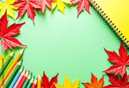 A frame of autumn yellow and red leaves, colored pencils, and a notebook on a green background. Back to school. Top view.
