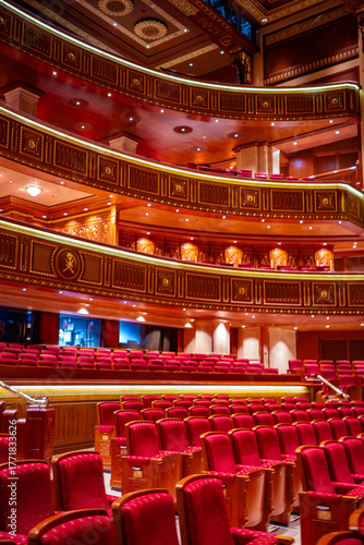 An elegant interior view of the Royal Opera House Muscat in Oman, featuring plush red velvet seats, golden wooden balconies of the luxurious design, and ornate architectural detailing