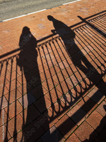 Shadows of two people cast on a brick pavement near a fence. The scene captures a moment of interaction in an urban setting.