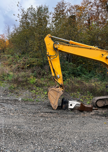 A yellow excavator scoop is parked on a gravel surface surrounded by trees and shrubs. The scene is set in an outdoor construction area