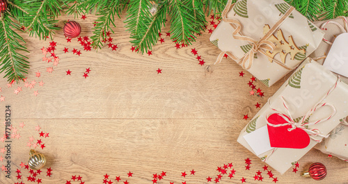 Christmas gifts wrapped in eco paper with fir branches, red confetti stars, and festive ornaments on wooden background, top view with copy space for text or greeting card design