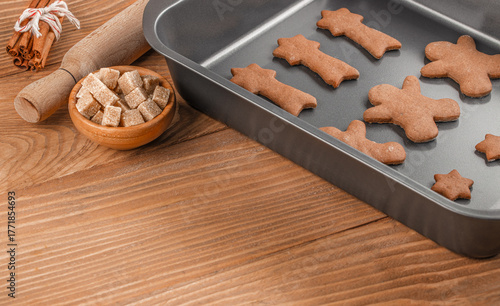 Raw gingerbread cookies on baking tray with brown sugar cubes, cinnamon sticks and rolling pin on rustic wooden table, preparation for Christmas baking, copy space