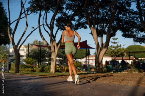Fit athletic woman doing cardio training outdoors, captured mid-jump with her back to the camera, wearing green shorts and sneakers, exercising on a park path in soft morning sunlight