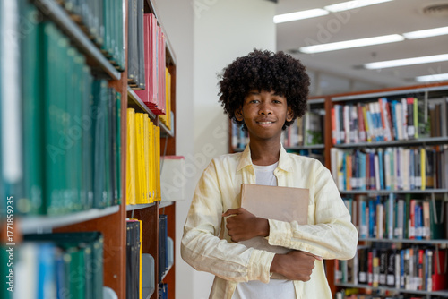 Obraz na płótnie Young male student standing in library, confidently holding book and smiling in educational school