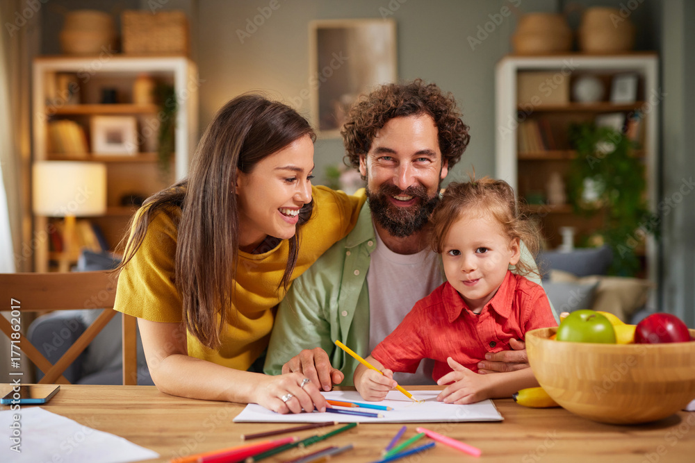 Fototapeta premium A happy family engages in a fun drawing activity at home. The mother and father smile at their young child who is coloring on a sheet of paper. Colorful art supplies are spread across the table.