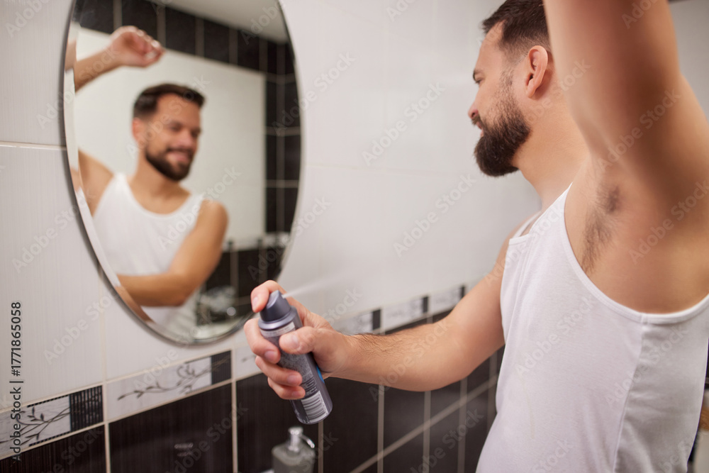 Obraz premium A man stands in front of a mirror, applying deodorant in a bathroom. He wears a tank top and smiles as he gets ready for his day.