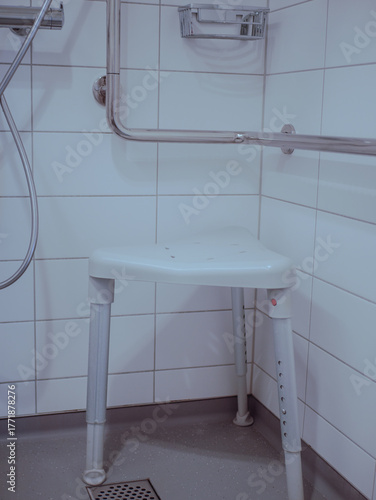 A medical shower chair placed in a hospital or rehabilitation clinic bathroom with tiled walls and stainless steel handrails. This hygiene equipment is designed to assist elderly or disabled patients