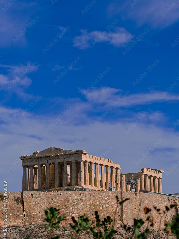 Obraz premium Parthenon, monumental symbol of Western civilization, stands atop the fortified Acropolis against a blue sky scattered with clouds, emphasizing the historical significance of the ancient structure.