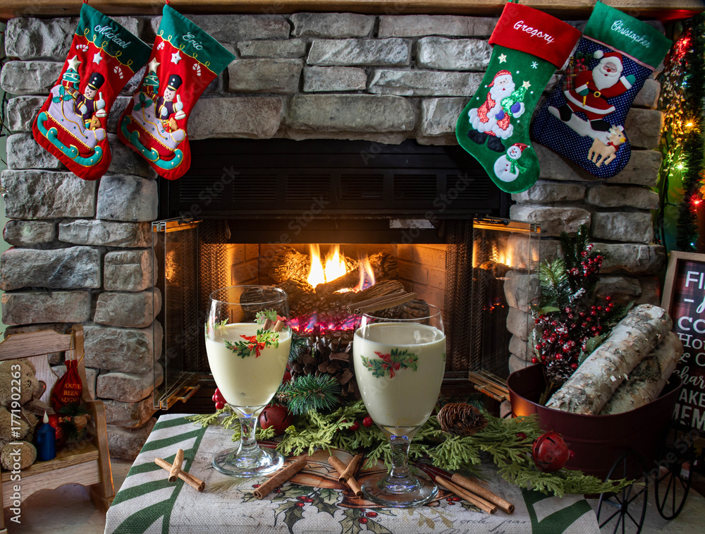 Obraz premium Two glasses of creamy holiday drinks sit on a table near a warm fireplace. Christmas stockings hang above, and decorations create a cozy atmosphere filled with cheer during the festive season.