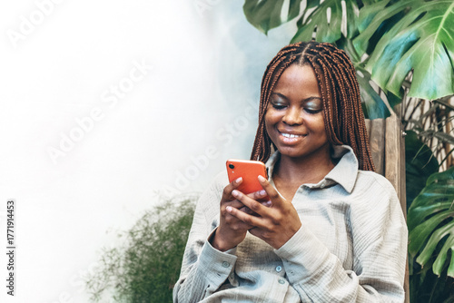 A cheerful woman with braided hair holds her smartphone in her hands surrounded by bright green nature