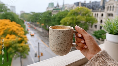 A woman's hand holding a coffee mug on a rainy day. Cozy autumn scene with a view of a city street from a balcony