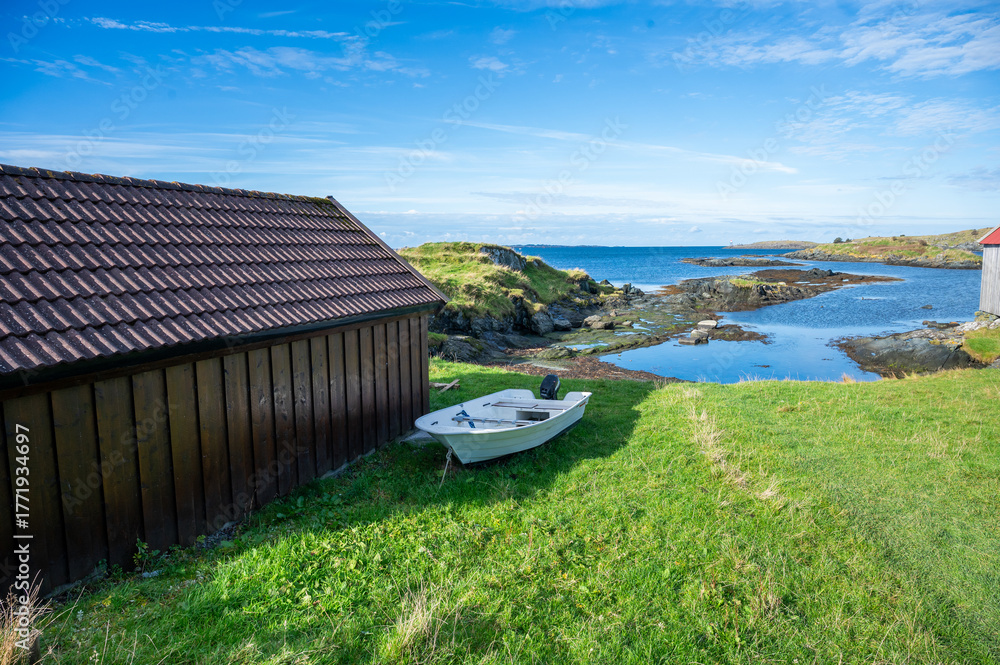 Fototapeta premium Serene coastal scene features a wooden shed and a small white boat on a grassy slope, with a clear blue sky and calm sea in the background, Haugesund, Norway