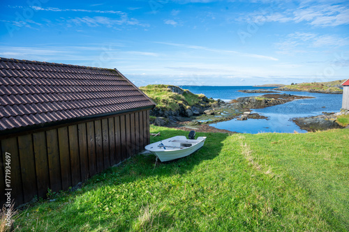 Serene coastal scene features a wooden shed and a small white boat on a grassy slope, with a clear blue sky and calm sea in the background, Haugesund, Norway