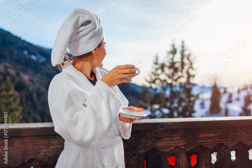 Young woman drinking coffee on hotel balcony enjoying winter mountains view wearing white robe and towel on head. Space