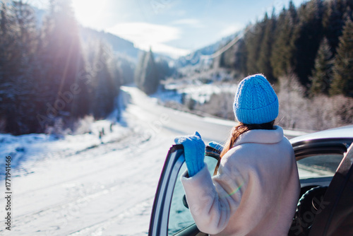 Back view of woman traveler admiring snowy mountains walking out of car looking at winter landscape. Traveling by auto