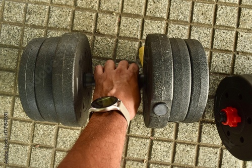 Adjustable Dumbbells on Outdoor Tile Floor in Park Setting
A pair of adjustable dumbbells placed on a tiled surface in an open-air park environment. The black weight plates are secured with red lock