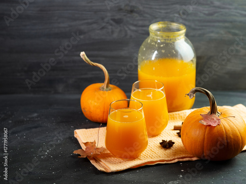 Delicious and healthy homemade pumpkin juice in a glass jar and glasses. Ripe orange pumpkins and spices. Dark background.