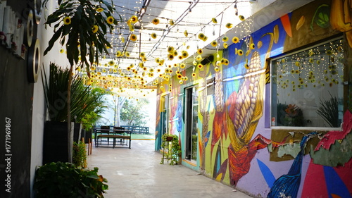 Pasillo decorado con girasoles colgantes y luces cálidas, creando un ambiente alegre y acogedor. Al fondo se observan murales coloridos y vegetación