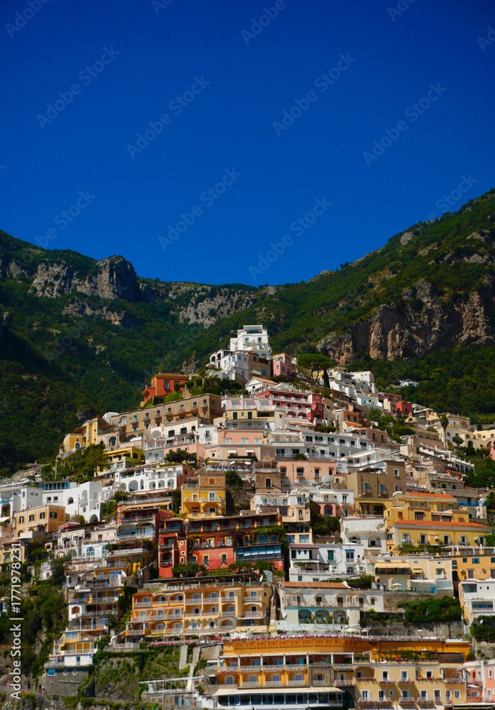 Naklejka premium Colorful hillside houses stacked above the blue waters of the Amalfi Coast in Positano, Italy, framed by steep green cliffs and a vivid blue summer sky on a clear sunny day.