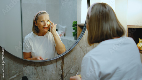 Elderly woman applying skincare in a well-lit bathroom while smiling in front of a round mirror during the day