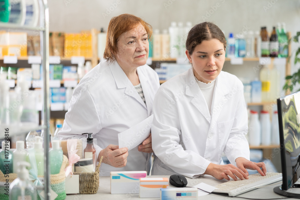 Fototapeta premium Elderly and young female pharmacists checking medical prescription on computer in pharmacy