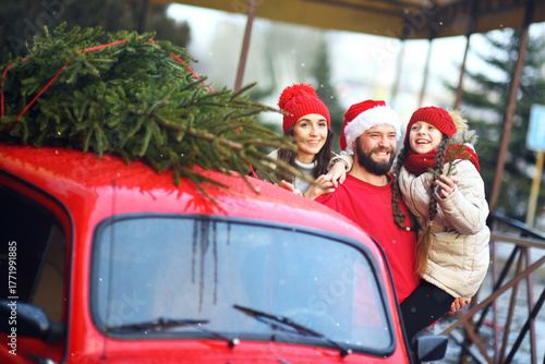mom, dad and a daughter in a knitted red hat with pigtails hug and smile against the background of a red car with a Christmas tree on the roof