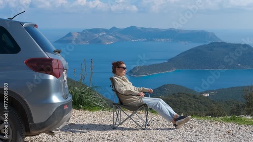 Tourist relaxing in folding chair enjoying island view. Resting woman enjoying breathtaking seascape from folding chair beside parked vehicle during scenic coastal road trip, experiencing freedom