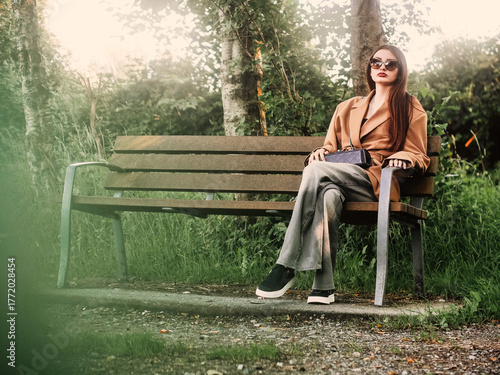 Young and stylish teenager girl sitting on a bench in a park alone. Waiting for date concept. The model wears trendy brown jacket and dark glasses and looks calm and elegant. Selective focus.