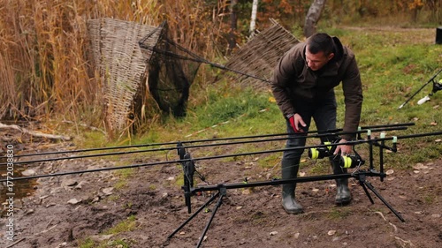 Fisherman setting up fishing rods on tripod near lake, man preparing tackle and adjusting reels on autumn day, outdoor hobby and recreation scene with natural background and reeds