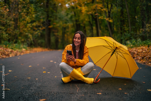 Smiling young woman in yellow raincoat and boots sits on autumn forest road with yellow umbrella, using smartphone and enjoying peaceful outdoor moment during fall season.