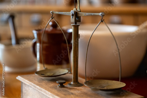 An antique brass two-plate balance scale in a historic Apothecary or pharmacy. There's a chemistry mortar and pestle with a club-shaped grinder used for medicine, vintage bowls, and old bottles. 