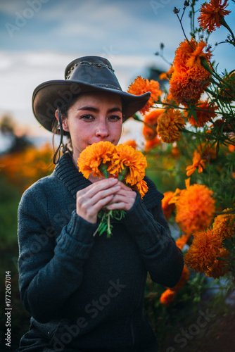 Beautiful girl in a cowboy hat smelling marigolds and looking at the camera in a field of marigold flowers that will be used for the traditional Day of the Dead festivities in Mexico
