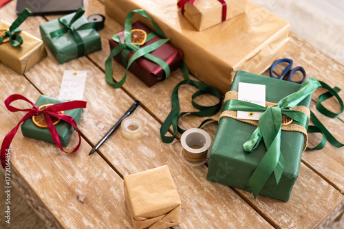 Green-wrapped box resting on rustic table, ribbons, tags, tape, twine and scissors lying scattered