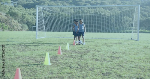 Practicing three boys wearing blue tops kicking soccer ball toward goal on valley pitch, with cones