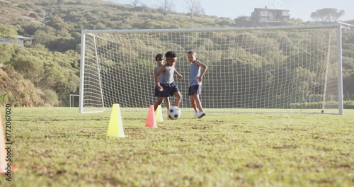 Three boys practicing soccer drills in grassy field, with ball and cones near goal