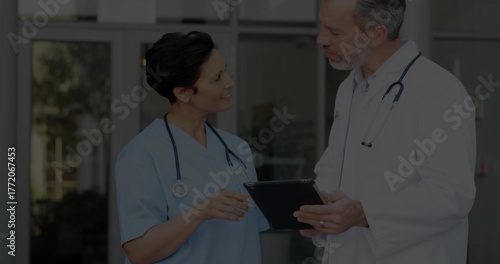 Doctor in lab coat and nurse in blue scrubs discussing at hospital doors, with tablet, stethoscopes