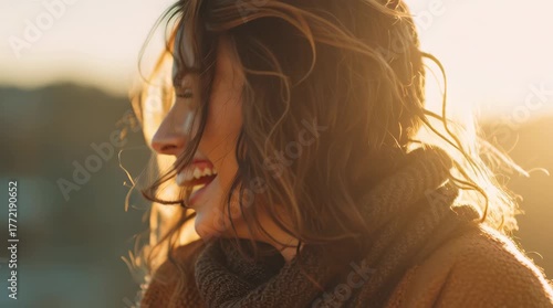 The woman has long, wind-swept brown hair and is wearing a chunky brown sweater or scarf. She first looks directly at the camera with a wide, genuine smile before turning her head to the side.