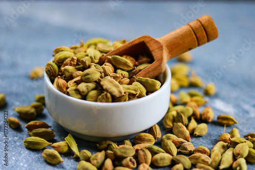 Heap of green cardamom seeds in bowl with wooden spoon on blue background