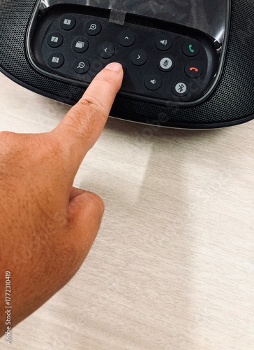 person’s hand pressing a button on an office conference speakerphone placed on a light wooden desk