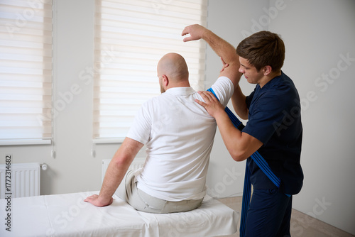 Chiropractor adjusting male patient’s shoulder in clinic. Physiotherapist performing manual therapy to relieve pain, improve mobility, support rehabilitation after injury or musculoskeletal disorder.